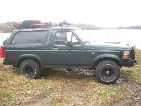 1993 Ford Bronco - A green Ford Bronco is parked on a muddy bank next to a body of water, equipped with a roof rack and off-road tires.