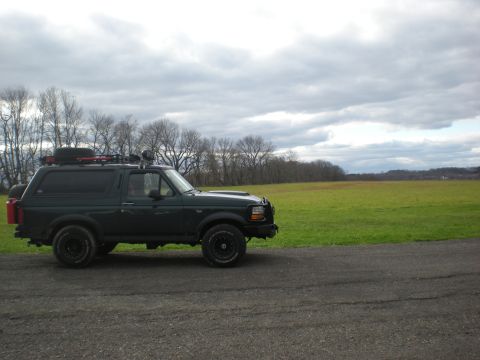 1993 Ford Bronco - A dark green 1993 Ford Bronco is parked on a gravel road with a large grassy field and trees in the background under a cloudy sky.