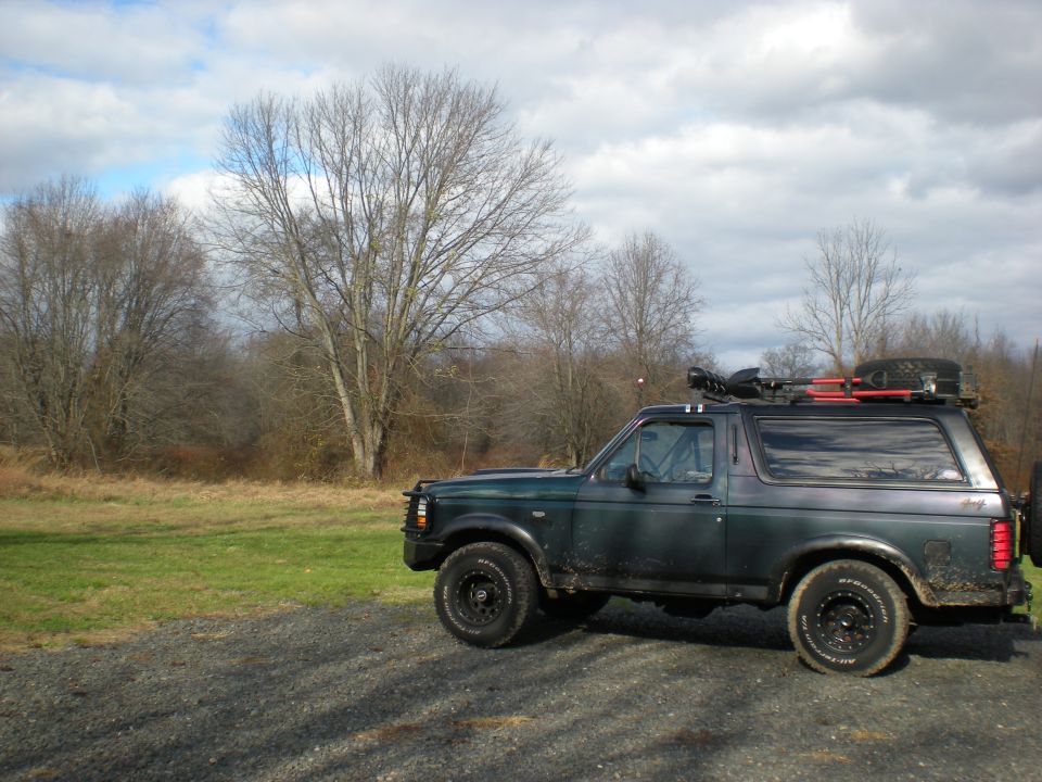 Dark green Ford Bronco parked on gravel, with roof rack, spare tire, and shovel.