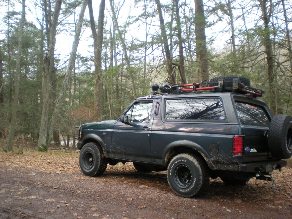 1993 Ford Bronco parked on a dirt road in a forest, with a roof rack and spare tire.