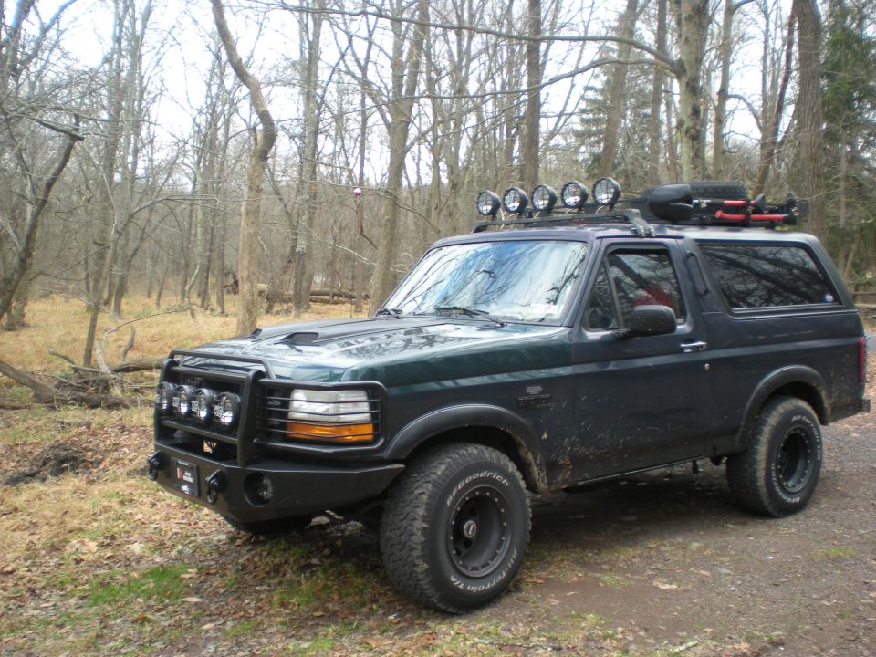 1993 Ford Bronco with roof rack lights, spare tire, and custom bumper in a wooded area.