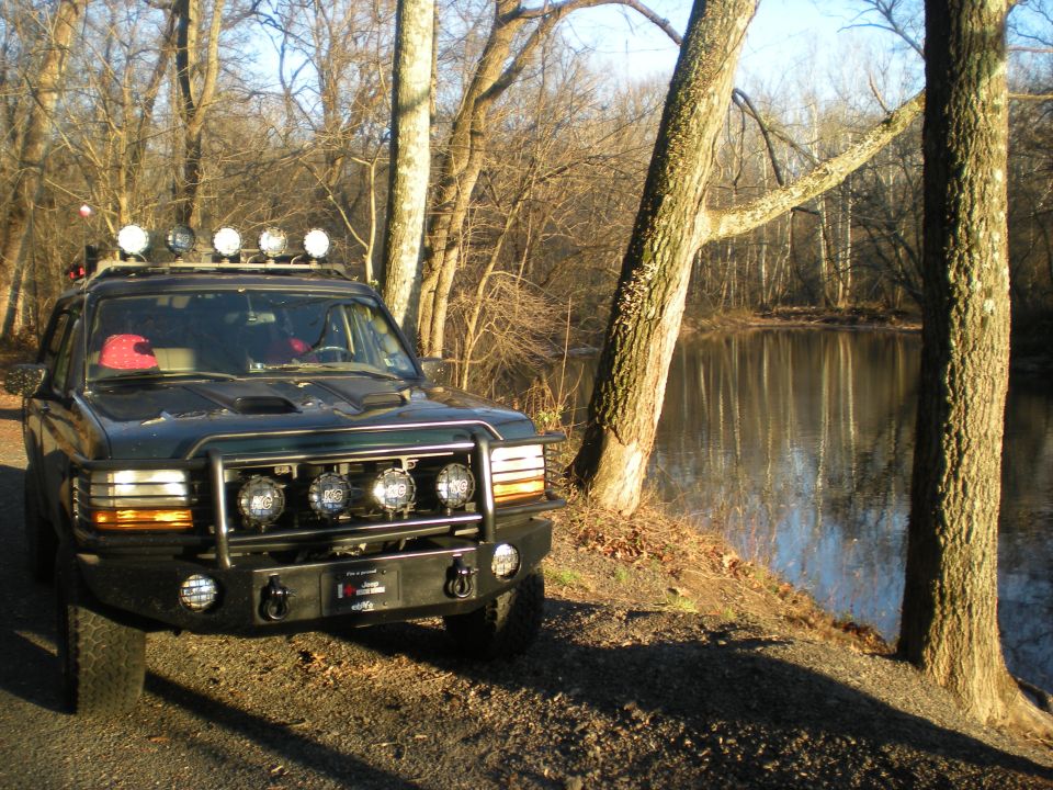 Dark green Ford Bronco with roof rack lights and brush guard lights parked by a lake.