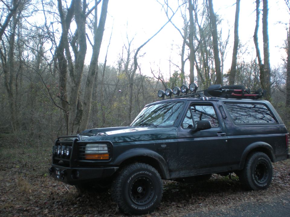Dark Ford Bronco with roof rack and off-road tires parked in a forest.