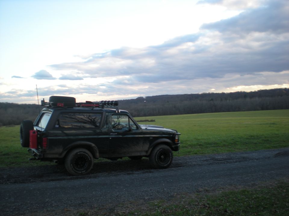 Dark green Ford Bronco with roof rack and spare tire parked on gravel road, green field and treeline in background.