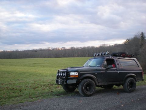 1993 Ford Bronco - A black SUV with a custom bumper and roof lights sits in a rural landscape under a cloudy sky.