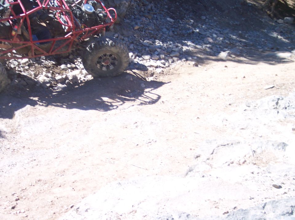 Red off-road buggy rear tire and suspension on a dirt and rocky trail.