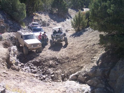 1995 Ford Bronco - Three off-road vehicles are parked on a rocky, dirt trail surrounded by trees. A tan SUV is in the foreground, with a white Jeep and a roll…