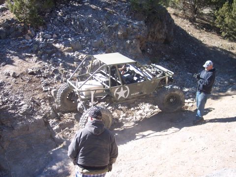 1995 Ford Bronco - A custom-built, olive green off-road buggy with a roll cage is navigating a rocky incline. Two men are visible, one standing near the buggy…