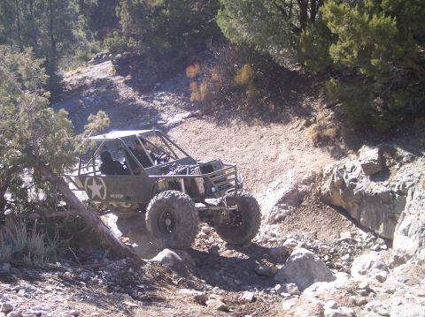 1995 Ford Bronco - A custom-built, olive green off-road buggy with large tires navigates a rocky, uphill trail surrounded by trees and brush.