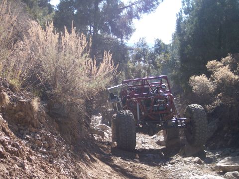 1995 Ford Bronco - A custom-built, dark red off-road buggy with large tires navigates a rocky, uphill trail surrounded by dry brush and trees.