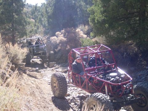 1995 Ford Bronco - Two off-road buggies are driving on a rocky, dirt trail through a dry, wooded area. The buggy in the foreground is red and has its engine e…