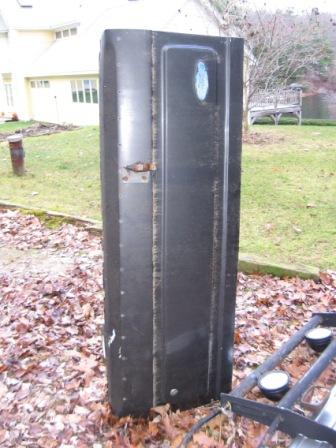 1993 Ford Bronco - A black SUV tailgate removed from the vehicle and stood on end in a leaf-covered yard, showing its outer skin with a rusting hinge or latch…