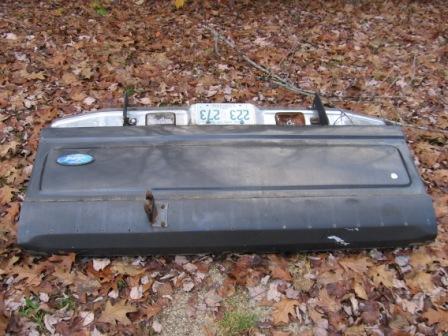 1993 Ford Bronco - A black tailgate lying face-up on a bed of fallen leaves, showing its top edge with a mounted license plate, plate light housings and a chr…
