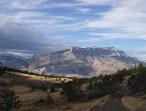 1990 Ford Bronco - A wide shot of a vast mountain landscape under a partly cloudy sky. A dirt road winds through the foreground, leading towards the distant,…