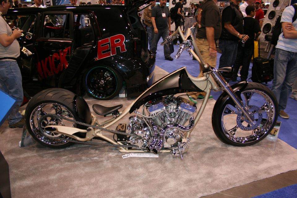 Custom chopper motorcycle with chrome engine and large rear tire, parked next to a black SUV with red lettering
