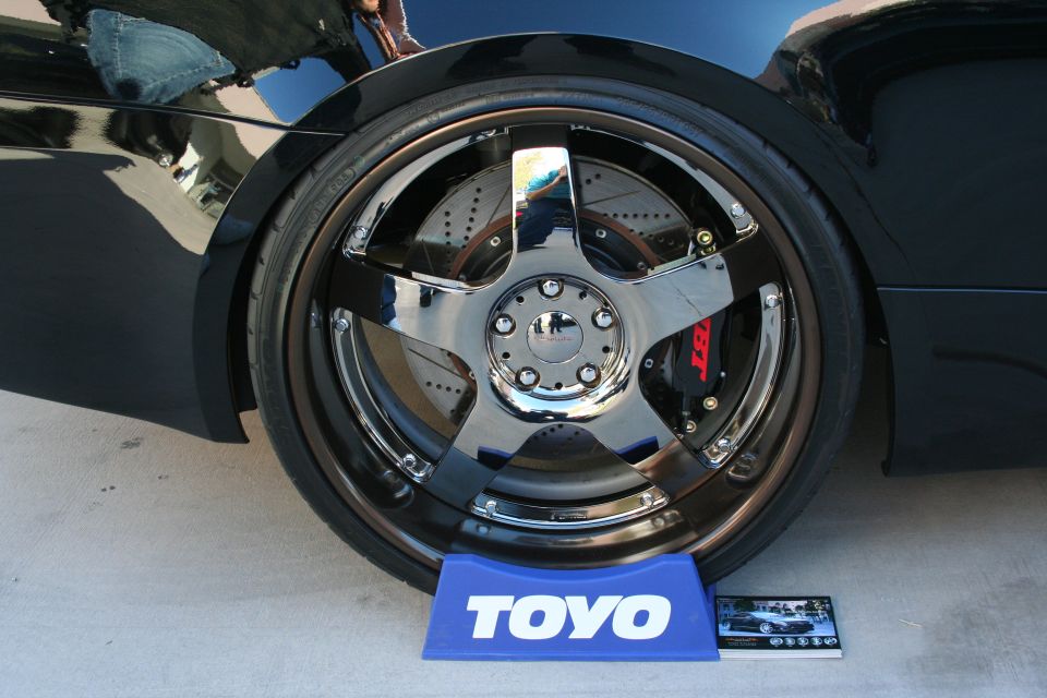 Black car's chrome wheel with red brake caliper, on a concrete surface with a TOYO tire stand