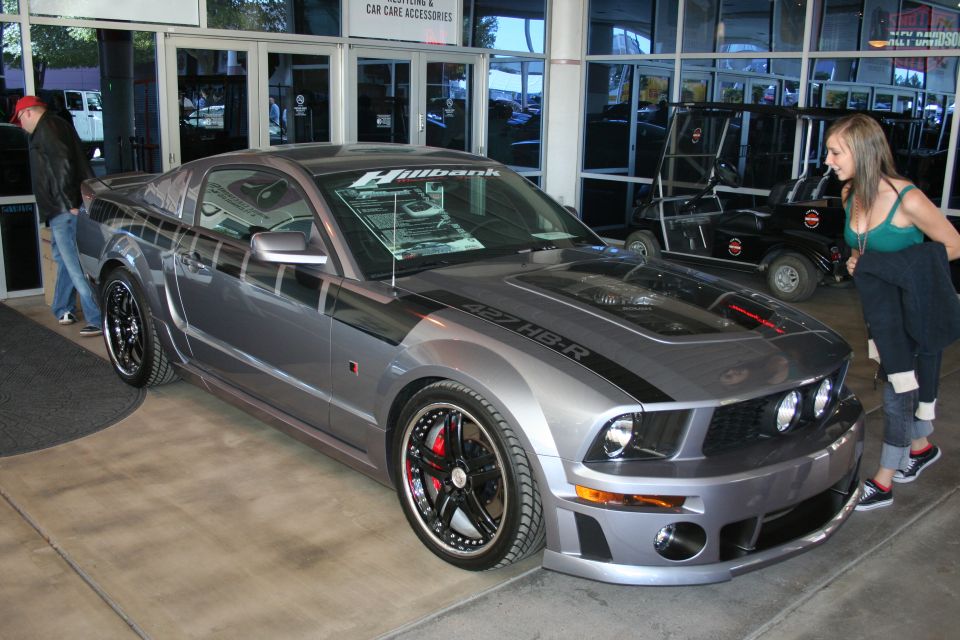 Gray Ford Mustang with black racing stripes and '427 HB-R' on the hood. A woman looks at it
