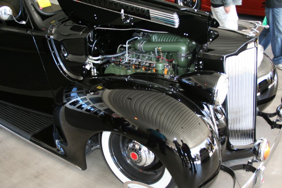 Black classic car with hood open showing a green supercharged engine and whitewall tires