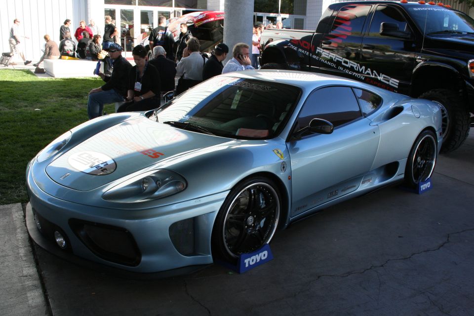 Light blue Ferrari coupe with racing decals and black wheels, parked next to a black pickup truck with "PERFORMANCE" text. TOYO brand blocks under wheels