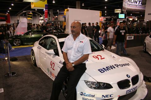 1998 Ford Crown Victoria - A man poses with a white BMW race car adorned with 'BMW Power' and '92' graphics at an auto show.