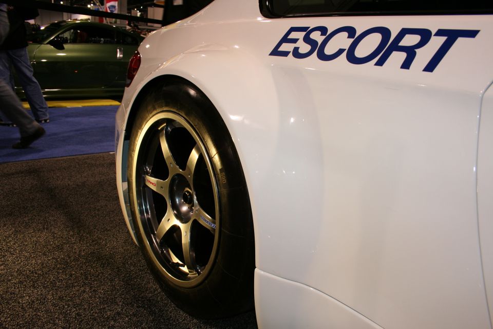 Close-up of a white race car's rear wheel and fender with blue ESCORT text. A green car is visible in the background