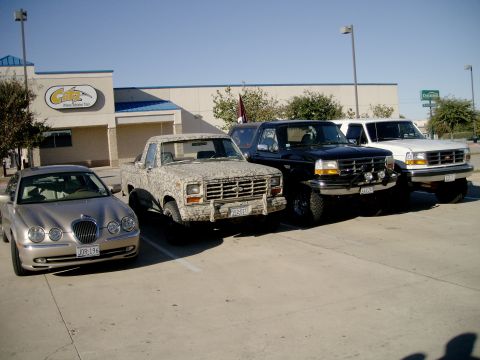 1984 Ford Bronco - A silver sedan, a camouflage pickup, a black SUV, and a white pickup are parked in a lot outside a building.