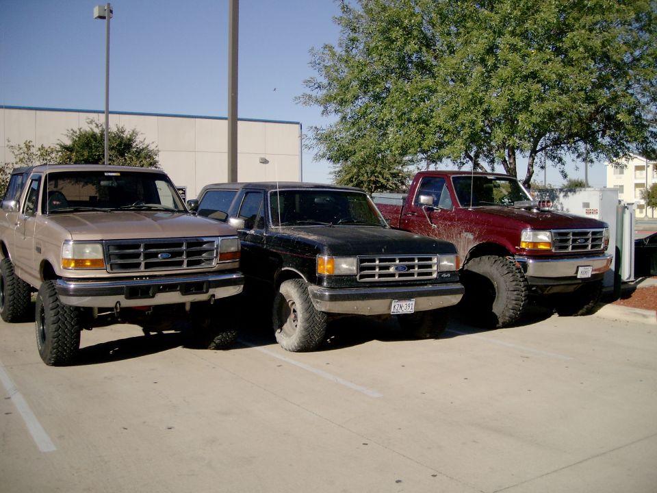 Three lifted Ford Broncos on mud tires, tan, black and red, parked in a lot