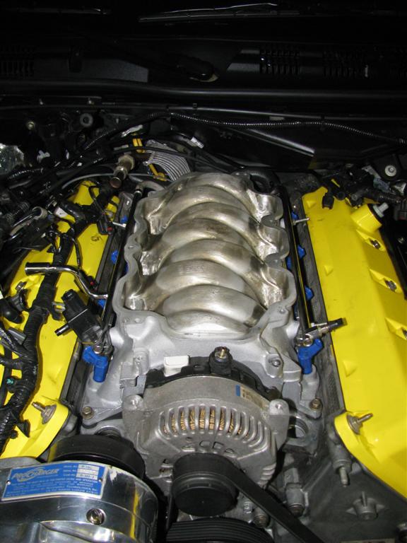 Engine bay with a polished intake manifold, yellow valve covers, and a ProCharger supercharger. Wires and hoses are visible