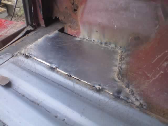Welded metal patch on a red vehicle's floor pan, showing rough welds and rust