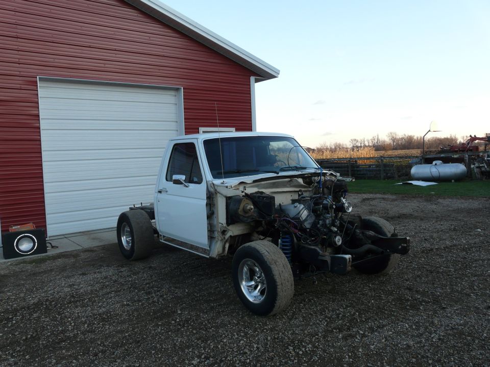 White Ford pickup stripped to cab and frame with exposed engine, parked by a red barn