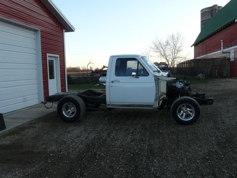 1994 Ford F-150 - Driver-side profile of a white Ford pickup cab and bare frame sitting on gravel beside a red barn, with the bed, front clip, fenders and ho…