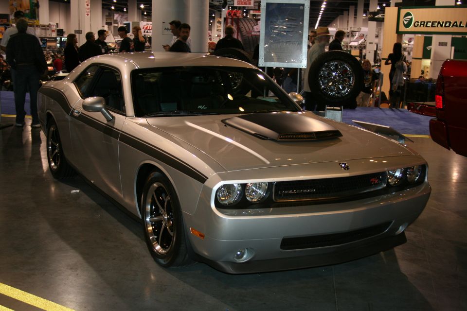 Silver Dodge Challenger with black racing stripes and hood scoop. It's at an indoor event with people in the background