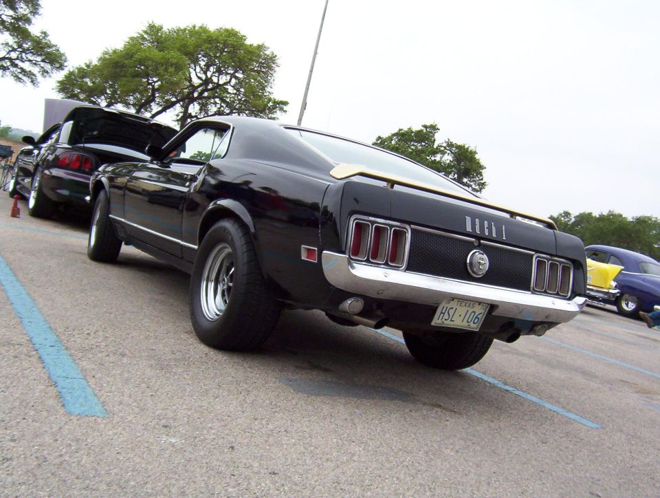 Black 1970 Ford Mustang Mach 1 parked, rear view. Another car with hood up is next to it