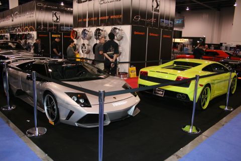 1998 Ford Crown Victoria - Two exotic sports cars are displayed at an automotive exhibition with various wheel brands in the background.