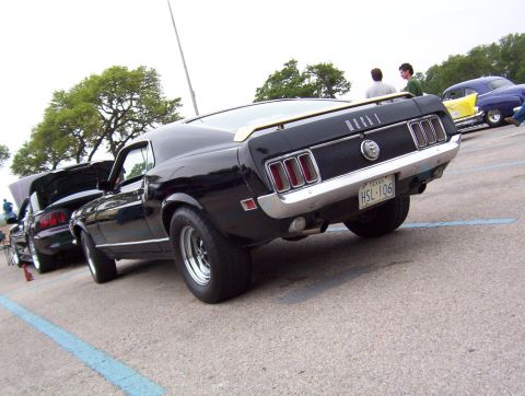 1970 Ford Mustang - A black 1969 Ford Mustang is parked next to another car with its hood up, with other vehicles in the background.