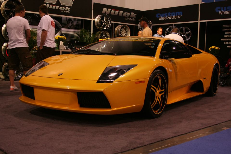 Yellow Lamborghini Gallardo on display at an auto show with custom wheels and people in the background
