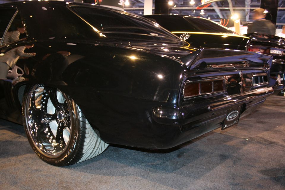 Rear quarter view of a black custom car with chrome wheels and a spoiler. Another black car is visible behind it