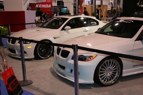Two white BMW coupes at an indoor event, one with black rims and the other with silver rims