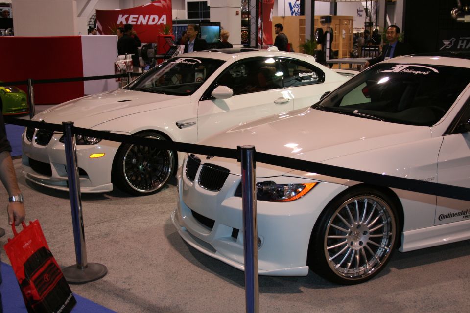 Two white BMW coupes at an indoor event, one with black rims and the other with silver rims