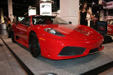 Red sports car with black wheels and racing stripes on the hood, parked indoors with a woman standing nearby
