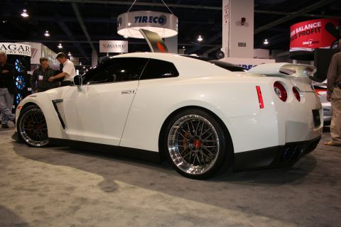 White sports car with custom wheels and a large rear spoiler, seen from the side at an indoor event
