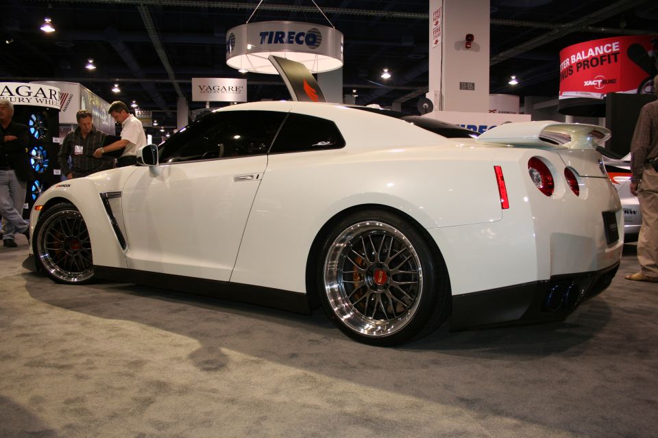 White sports car with custom wheels and a large rear spoiler, seen from the side at an indoor event