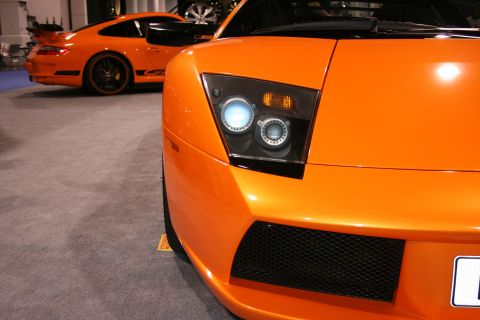 Orange sports car front end with distinctive headlights and grille. Another orange sports car is visible in th