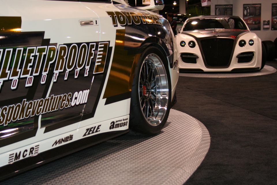 Close-up of a customized white car with 'BULLETPROOF AUTOMOTIVE' graphics, showing its side and wheel. Another white car is visible in the background