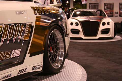 Close-up of a white and gold sports car's rear wheel, with another white car in the background
