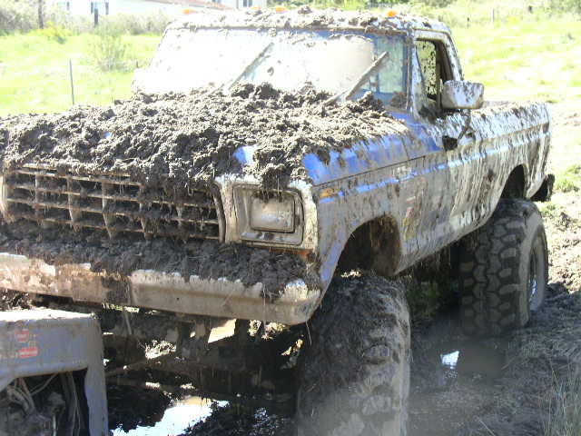 Mud-covered blue Ford pickup truck with large off-road tires stuck in mud.