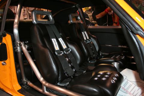 Car interior with black racing seats, five-point harnesses, and a roll cage. Orange exterior visible