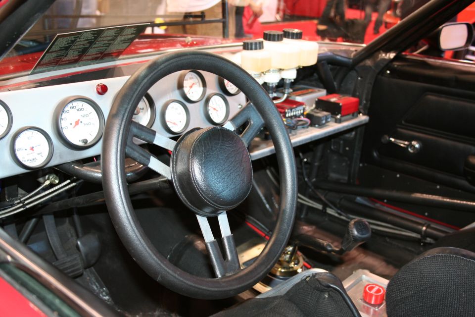 Interior of a race car with a large steering wheel, multiple gauges on the dash, and a roll cage