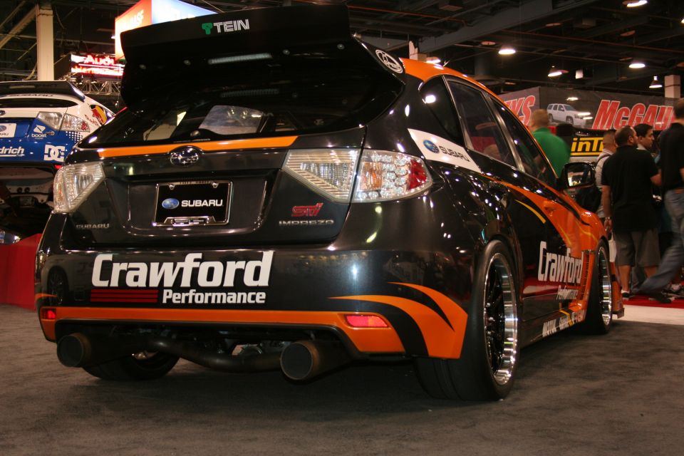 Rear view of a black and orange Subaru Impreza WRX STI hatchback with 'Crawford Performance' livery and a large rear spoiler. Another car is visible to the left