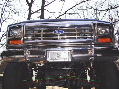 1986 Ford Bronco - Head-on view of a lifted dark-colored Ford truck with a polished chrome bar grille, blue Ford oval, rectangular headlights with amber corne…